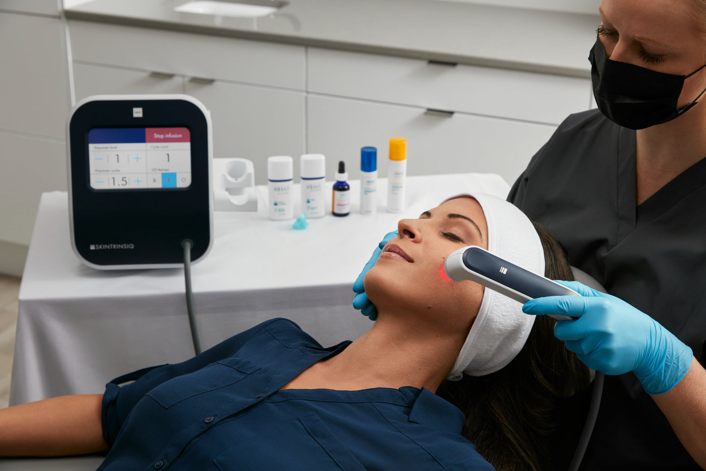 A skincare professional wearing gloves and a face mask performs a facial treatment on a woman using a handheld device emitting red light, with Obagi products and a Skintrinsiq machine visible in the background.
